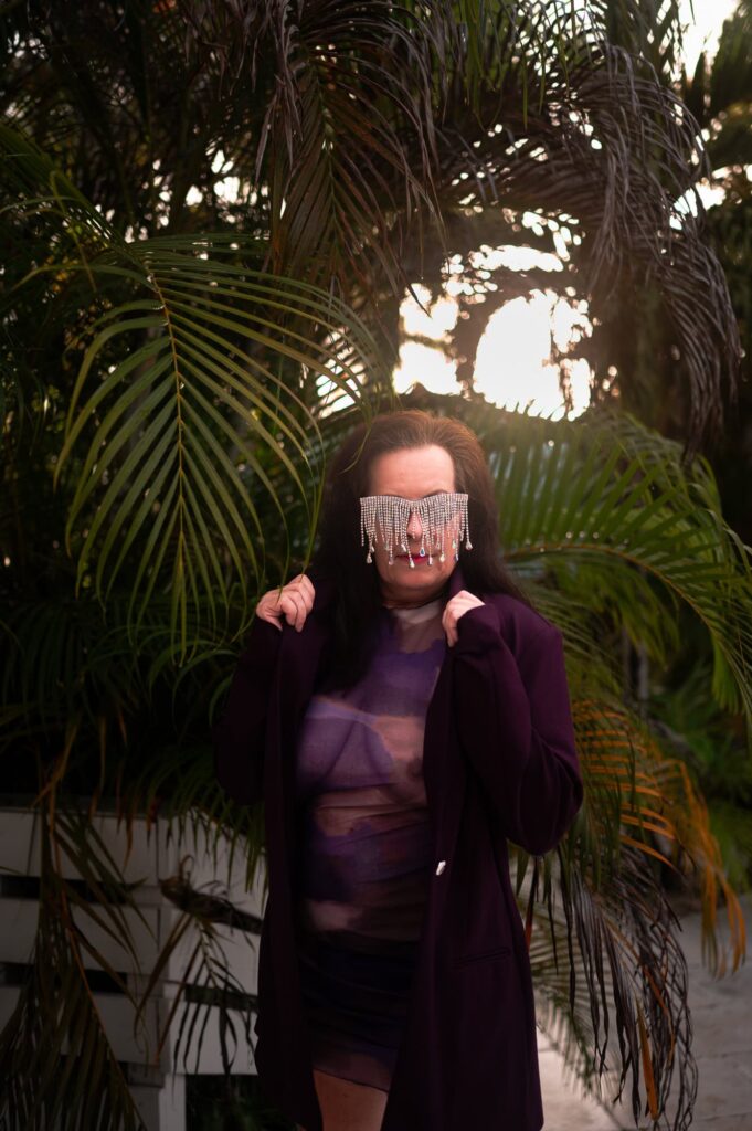 Boudoir photography for women over 40. This photograph is of a 60 year old woman wearing a purple dress and blazer. She is standing amongst some ferns. This photograph was taken by ELI Boudoir.