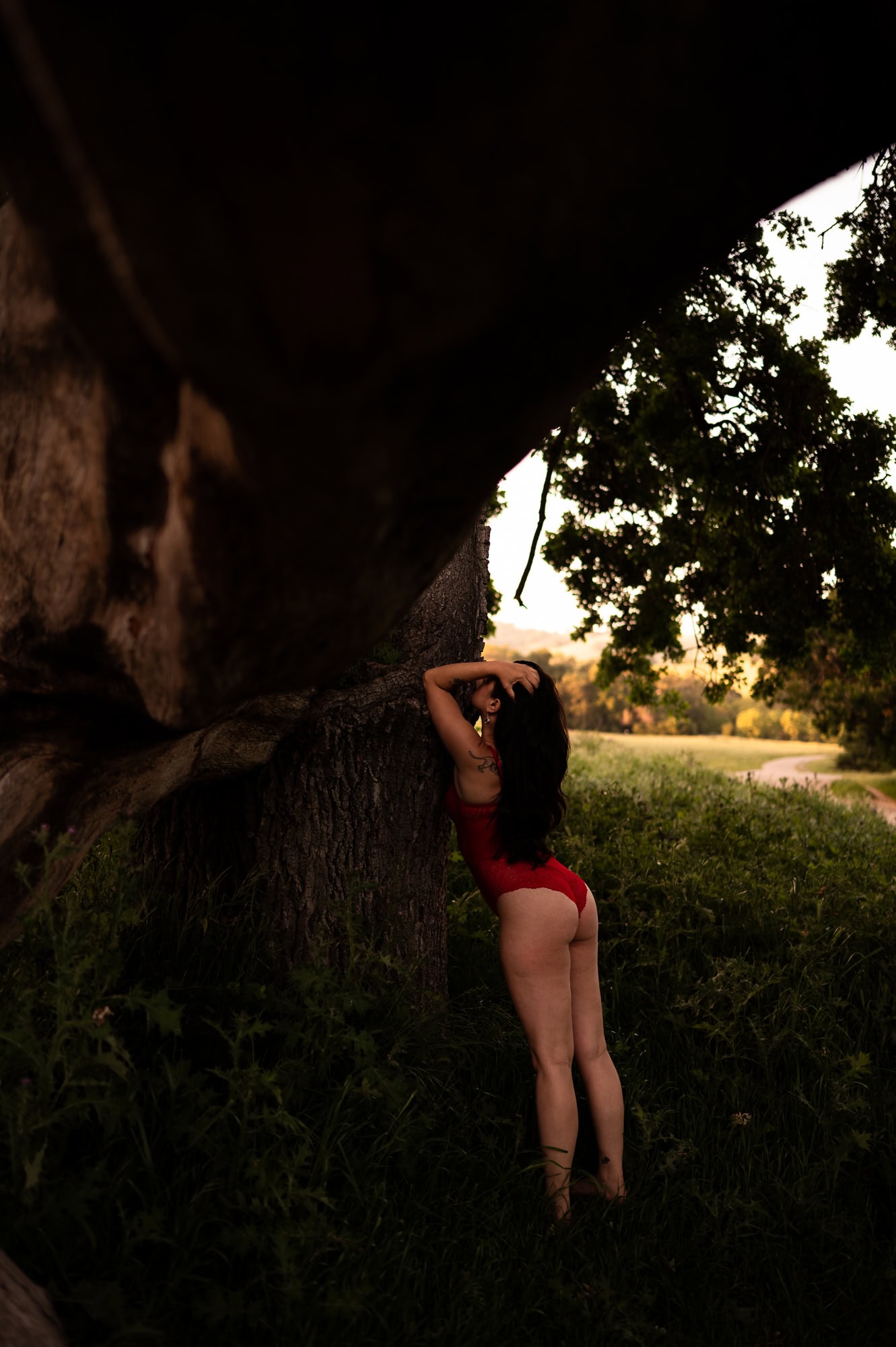 Boudoir photography for women over 40. This photograph is of 40 year old woman in a red bodysuit She is standing in a meadow and posing standing up against a tree. This photograph was taken by ELI Boudoir.
